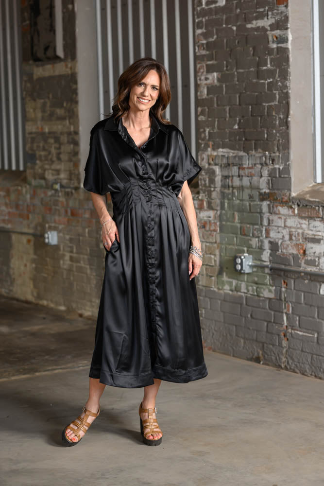 A woman wearing a black satin midi dress with a shawl collar and tie waist, standing in an industrial loft setting with brick walls.