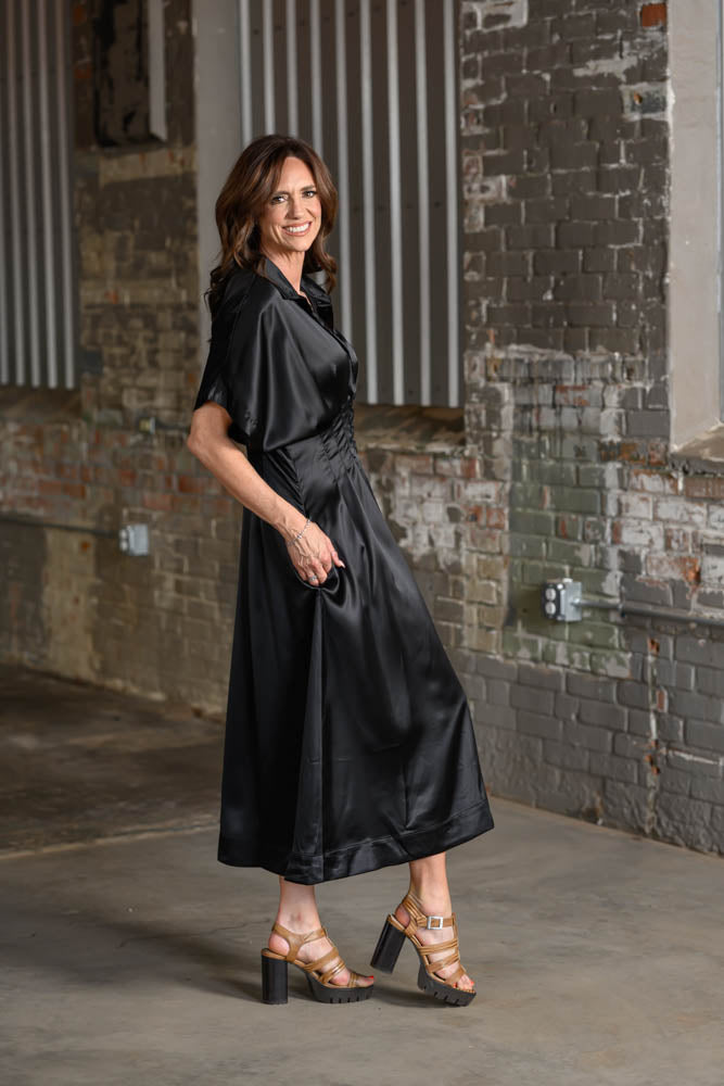 A woman wearing a black satin midi dress with a shawl collar and side pockets, paired with brown strappy high-heeled sandals, standing in an industrial brick alleyway.