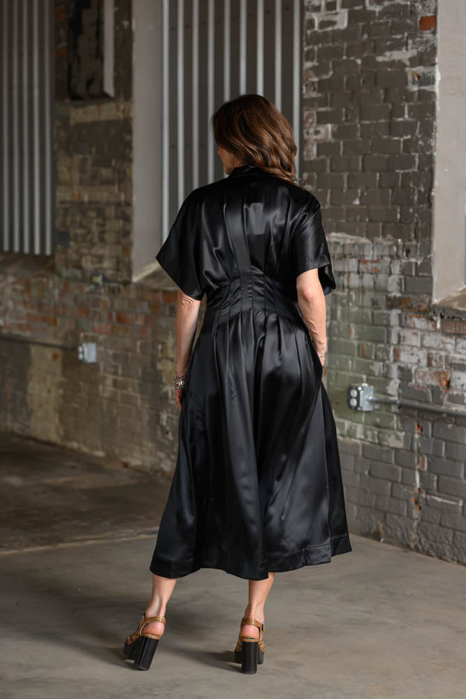 A woman viewed from behind wearing a black satin short-sleeve midi dress with a gathered waist, standing in an industrial brick alleyway.