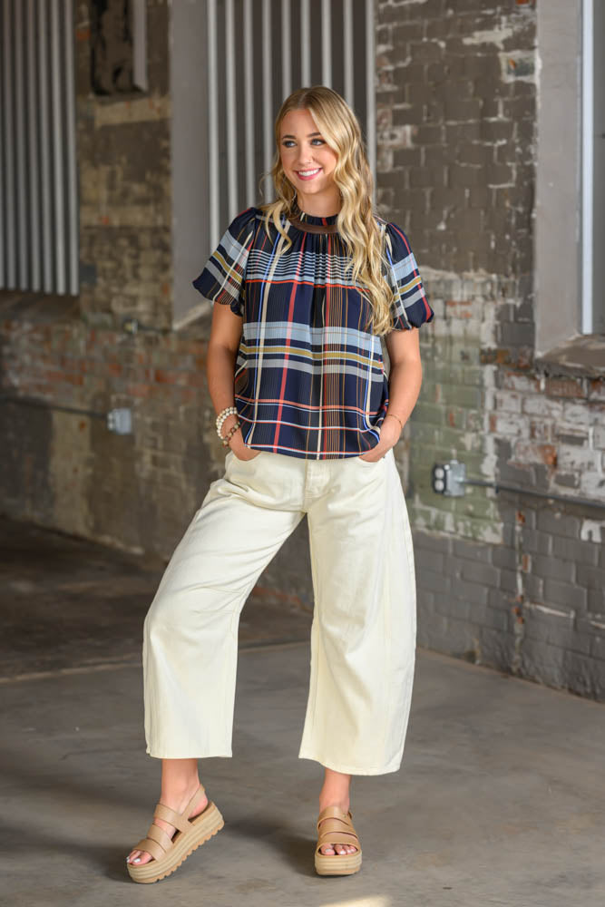 A woman modeling a short-sleeve plaid top with cream wide-leg cropped pants and tan wedge sandals, standing in front of a rustic brick wall.