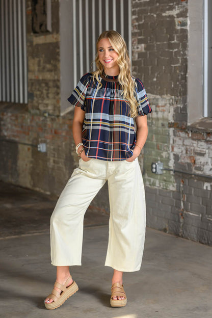 A woman modeling a short-sleeve plaid top with cream wide-leg cropped pants and tan wedge sandals, standing in front of a rustic brick wall.