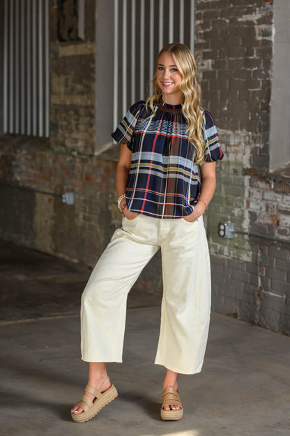 A woman modeling a short-sleeve plaid top with cream cropped wide-leg pants and tan platform sandals, standing in an industrial loft setting with a brick wall background.
