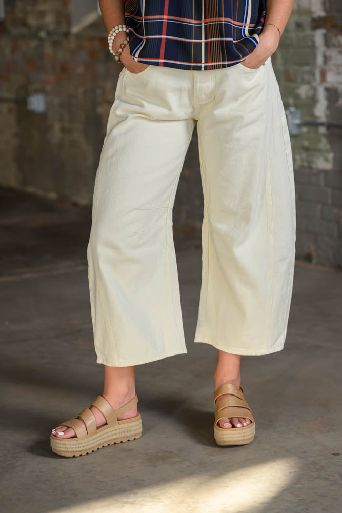 A person wearing cream-colored cropped wide-leg pants, a navy plaid top, and beige platform sandals, standing in a rustic indoor setting with a concrete floor and brick wall.