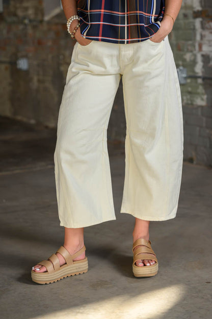 A person wearing cream-colored cropped wide-leg pants, a navy plaid top, and beige platform sandals, standing in a rustic indoor setting with a concrete floor and brick wall.