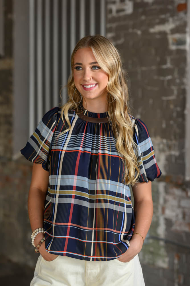 A smiling blonde woman wearing a short-sleeve plaid blouse with navy, red, yellow, and white stripes, paired with cream pants, standing against a rustic brick wall.