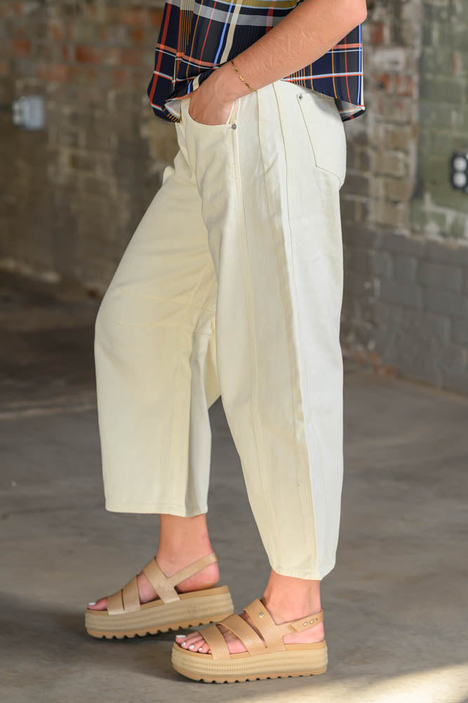 A person wearing cream-colored wide-leg cropped pants, a navy plaid shirt, and beige platform sandals, standing in front of a brick wall.