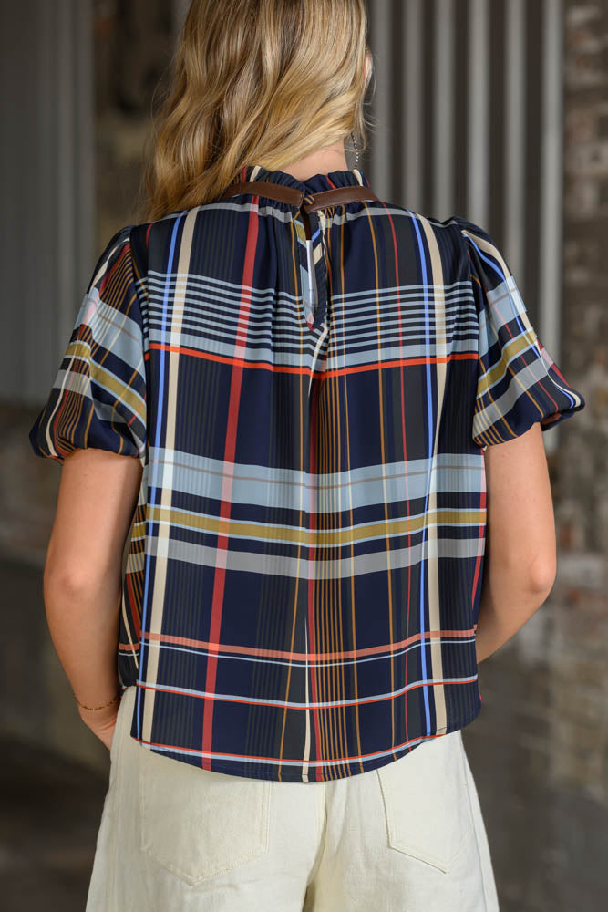 A woman viewed from behind wearing a short-sleeve plaid button-up blouse with navy, red, yellow, and white stripes, paired with light beige pants.