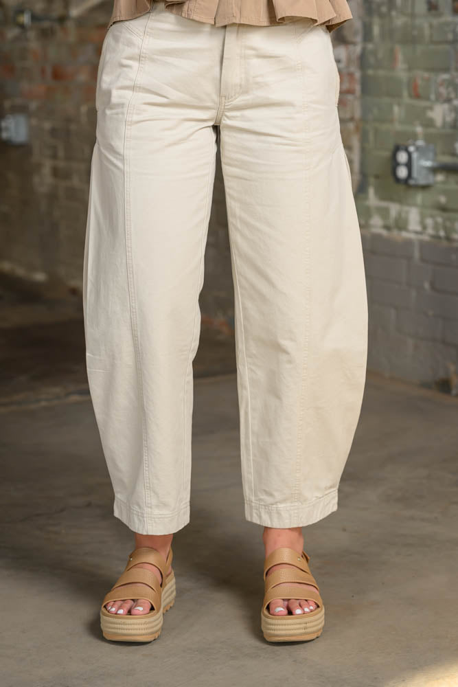 A person wearing loose, wide-leg cropped pants in a light beige or cream tone, paired with tan platform sandals, standing in an industrial interior with a brick wall and concrete floor.