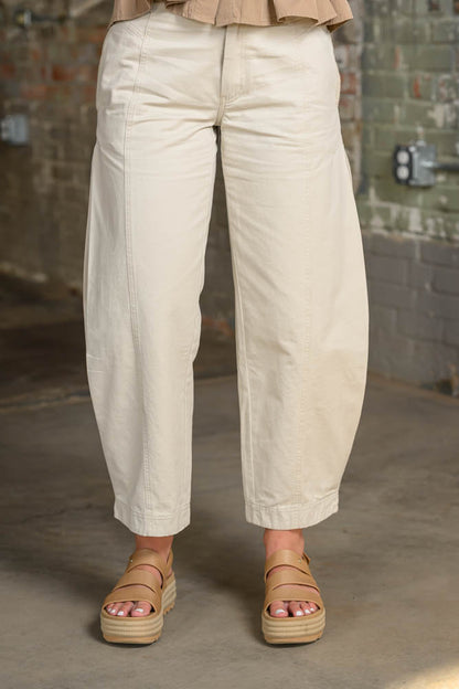 A person wearing loose, wide-leg cropped pants in a light beige or cream tone, paired with tan platform sandals, standing in an industrial interior with a brick wall and concrete floor.