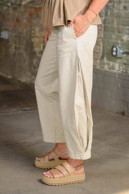 A person wearing loose, wide-leg cropped pants in a light beige or cream tone, paired with tan platform sandals and a matching beige top, standing in an indoor industrial setting with a brick and concrete wall background.