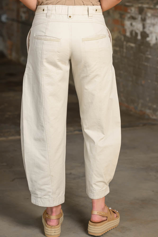 Back view of a person wearing cream-colored wide-leg cropped pants with a beige top and platform sandals, standing against a rustic brick wall.