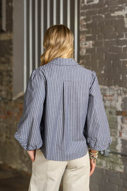 A woman viewed from behind wearing a blue and white vertically striped blouse with puffed balloon sleeves and beige pants, standing in front of a rustic brick wall.