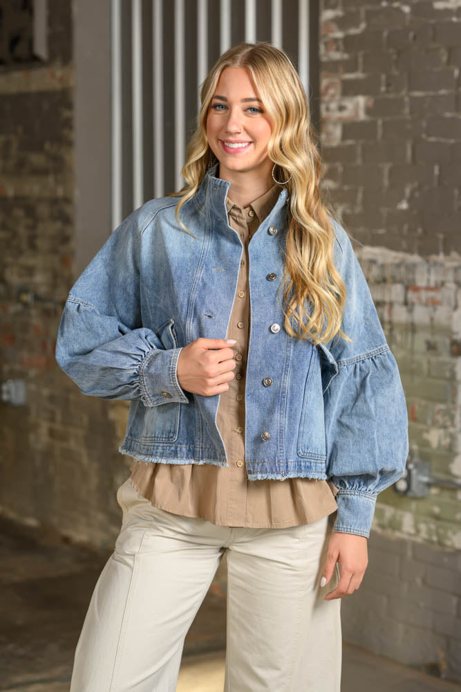 A woman modeling a light blue cropped denim jacket with dramatic puffed balloon sleeves, worn over a beige ruffled blouse and paired with cream pants, set against an industrial brick wall background.