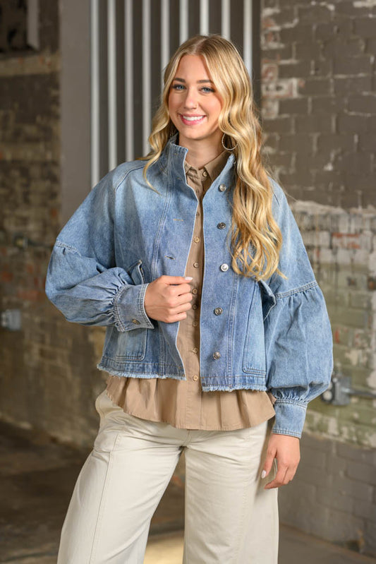 A woman modeling a light blue cropped denim jacket with dramatic puffed balloon sleeves, worn over a beige ruffled blouse and paired with cream pants, set against an industrial brick wall background.