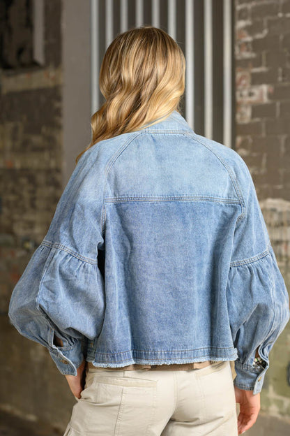 A woman viewed from behind wearing a cropped light blue denim jacket with dramatic balloon sleeves and beige pants, standing in front of a brick wall.