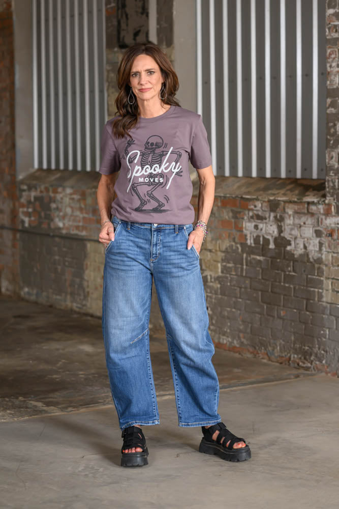 A woman stands in an industrial loft setting wearing a mauve-purple graphic t-shirt with a skeleton and the text 'Spooky Movies', paired with wide-leg blue jeans and black platform sandals.