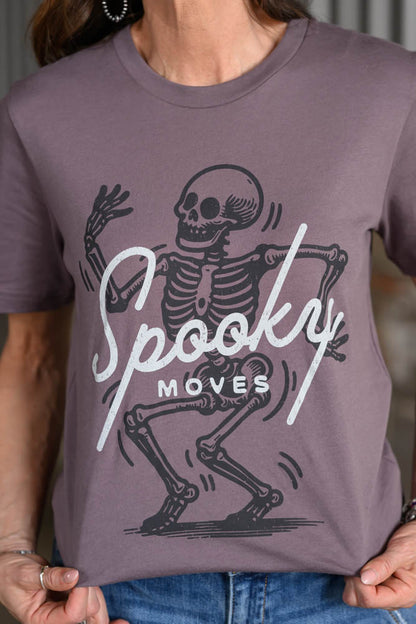 A person wearing a mauve-purple short-sleeve t-shirt with a black and white graphic of a dancing skeleton and the text 'Spooky Moves'.