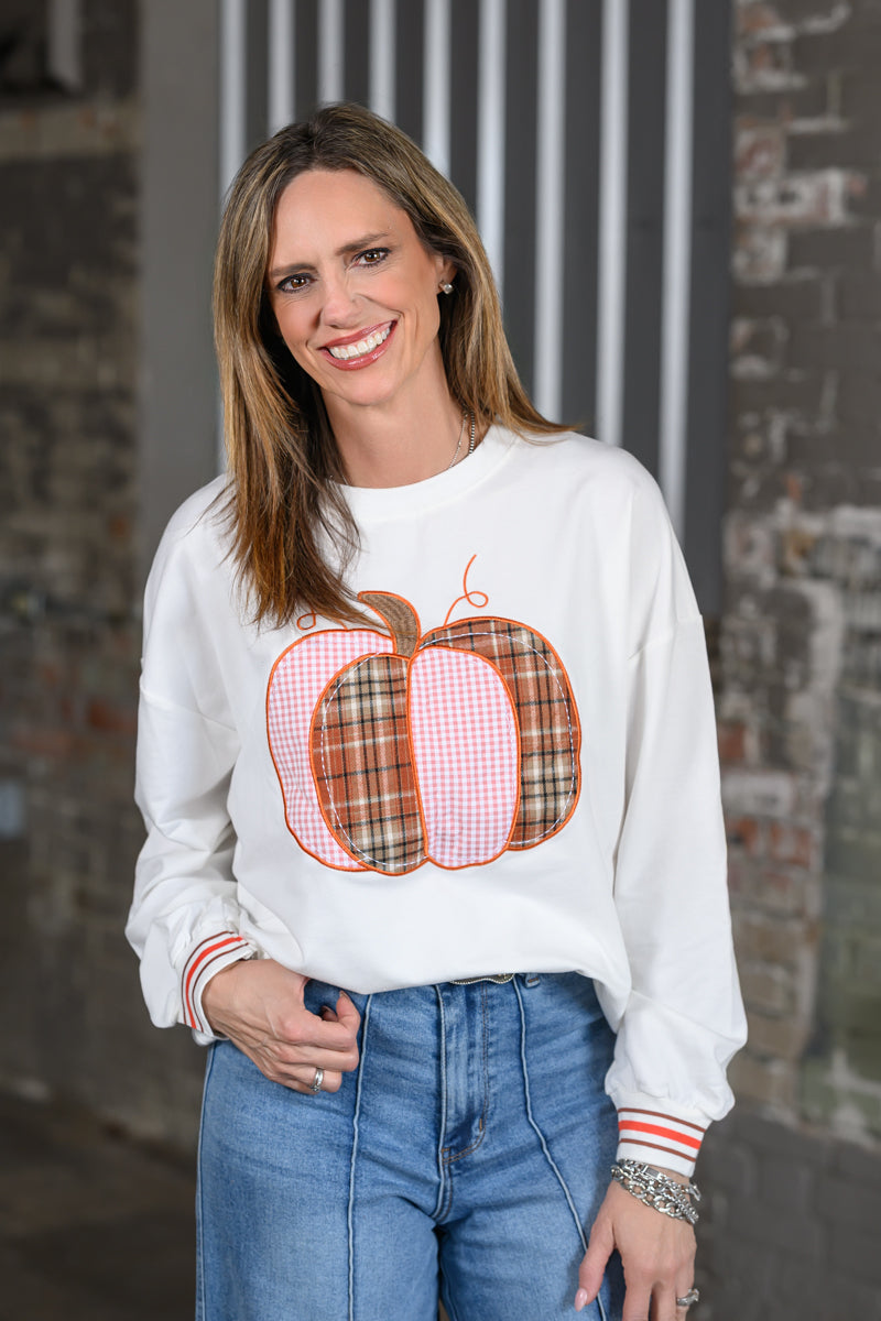A woman wearing a white crewneck sweatshirt with a large orange and red plaid pumpkin graphic on the front, paired with blue jeans, standing in front of a rustic brick wall.
