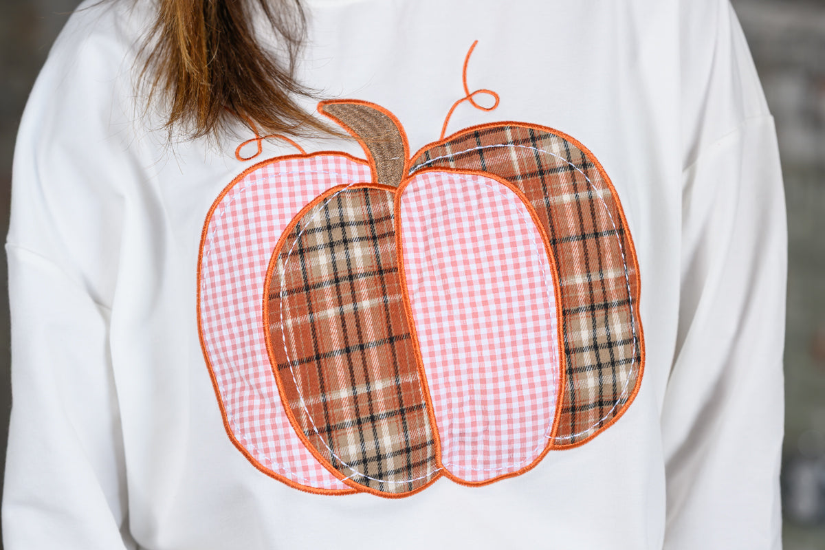 A close-up of a person wearing a white sweatshirt with a large applique pumpkin design on the front. The pumpkin features alternating pink gingham and brown plaid sections with orange stitching and a brown stem.