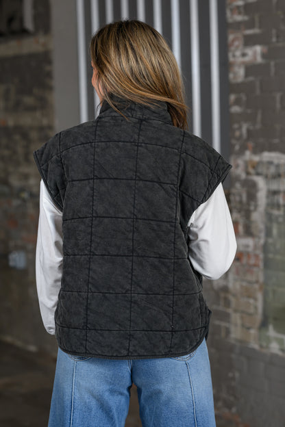 A woman viewed from behind wearing a dark charcoal gray quilted vest over a white long-sleeve shirt and blue jeans, standing in front of a rustic brick wall.