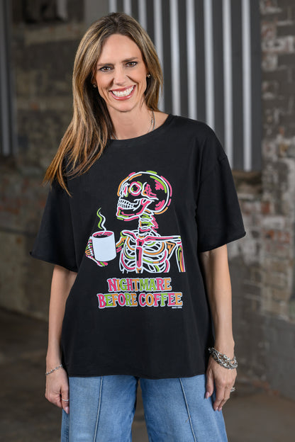 A woman wearing a black graphic t-shirt with a colorful skeleton holding a coffee mug and the text 'NIGHTMARE BEFORE COFFEE', paired with blue jeans, standing in front of a rustic brick wall.