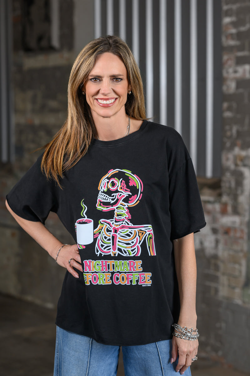 A woman wearing a black graphic t-shirt with a colorful skeleton holding a coffee cup and the text 'NIGHTTIME BEFORE COFFEE' in neon-style lettering, paired with blue jeans, standing against a rustic brick and metal wall.