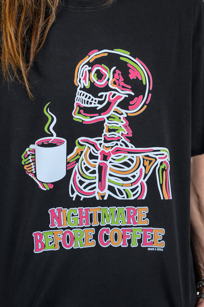A close-up of a black graphic t-shirt featuring a colorful cartoon skeleton holding a coffee mug with the text 'NIGHTMARE BEFORE COFFEE'. A person's hair and arms are visible at the edges of the frame.
