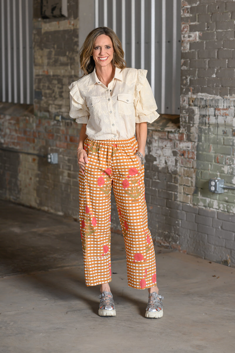A woman stands in an industrial loft setting with brick walls, wearing a cream puff-sleeve button-up shirt and orange-and-white gingham cropped pants with a coral floral print.