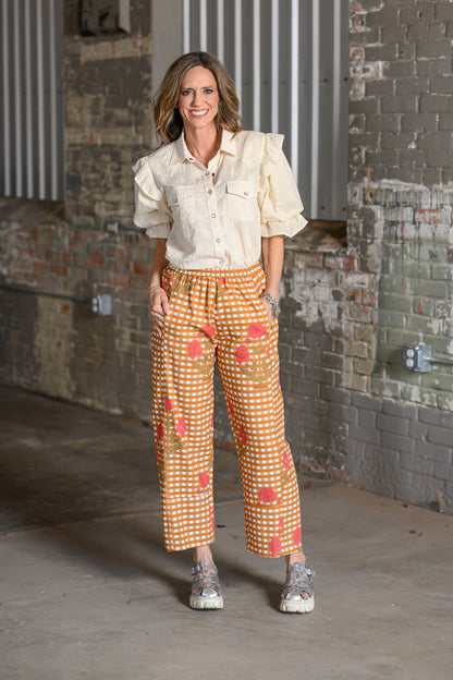 A woman stands in an industrial loft setting with brick walls, wearing a cream puff-sleeve button-up shirt and orange-and-white gingham cropped pants with a coral floral print.