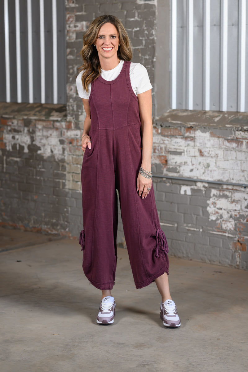 Jennifer Smith modeling a pair of wide-leg burgundy jumpsuit overalls over a white t-shirt, standing in front of a brick wall and corrugated metal building facade.