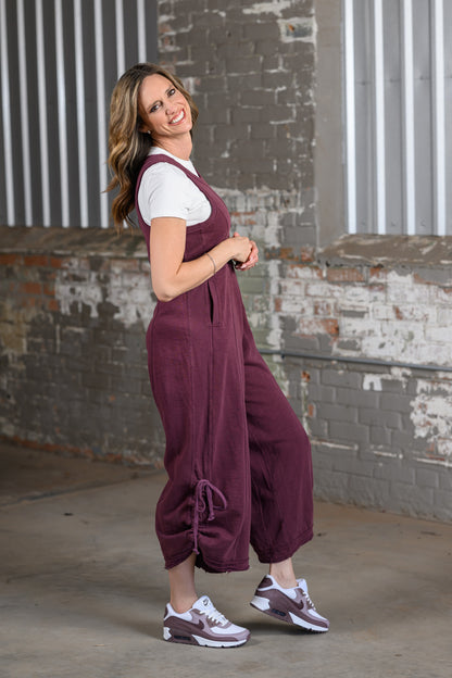 A woman modeling a pair of wide-leg corduroy overalls in a deep burgundy color, styled over a white t-shirt and sneakers, standing in front of a rustic brick wall.
