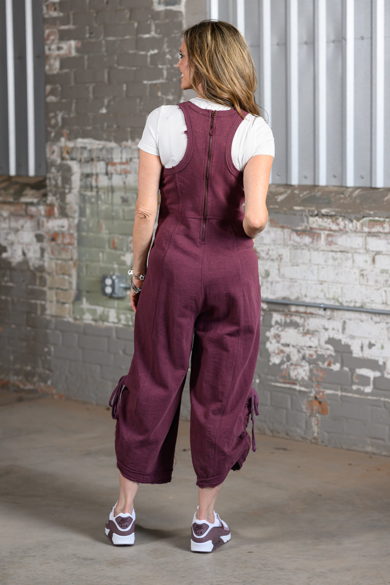 A woman viewed from behind wearing a maroon cropped overall jumpsuit over a white t-shirt, standing in front of a brick and corrugated metal wall.