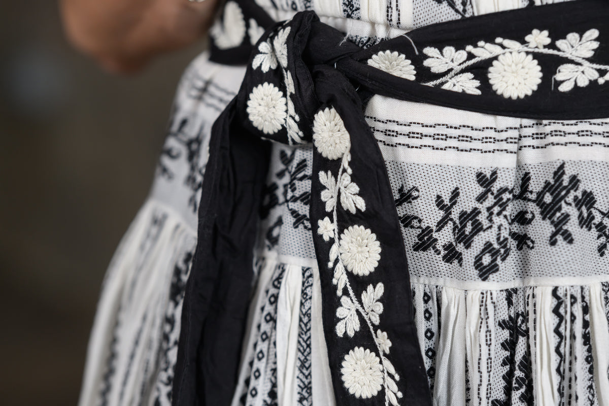 Close-up of a black and white embroidered traditional dress with a wide black sash tied at the waist, featuring white floral embroidery on a white woven fabric.