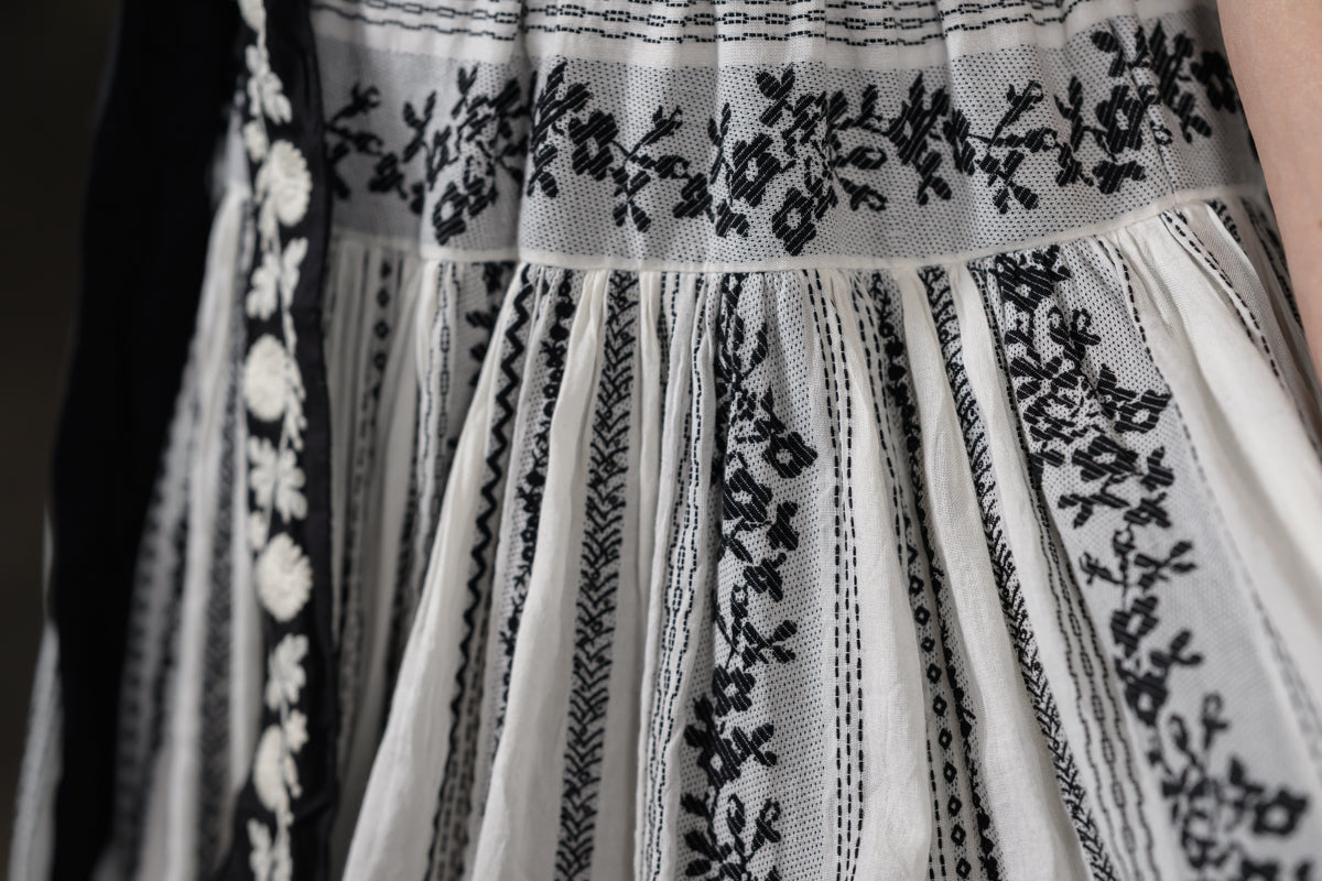 Close-up of a white embroidered dress with black floral embroidery and vertical gathered panels.