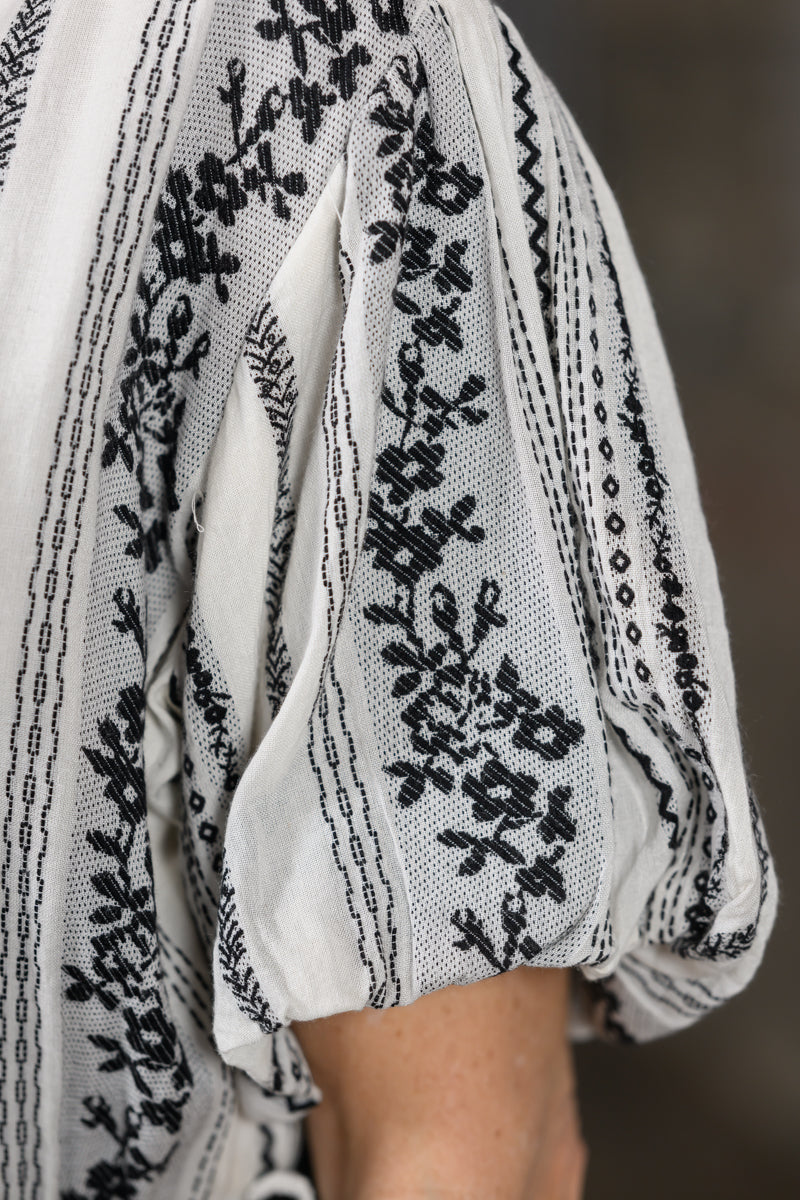 Close-up of a white or off-white sheer embroidered dress with black floral and geometric embroidery, worn with a matching black-and-white skirt or dress.