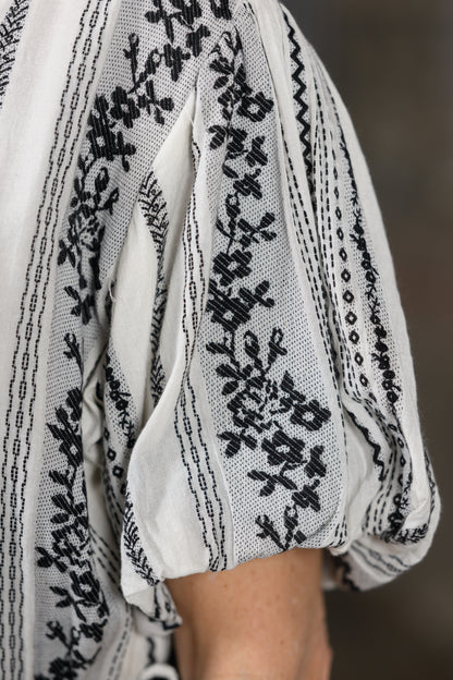 Close-up of a white or off-white sheer embroidered dress with black floral and geometric embroidery, worn with a matching black-and-white skirt or dress.