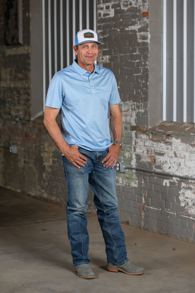 A man wearing a light blue polo shirt, blue jeans, gray suede boots, and a white cap with a brown logo, standing in front of a brick and corrugated metal wall.