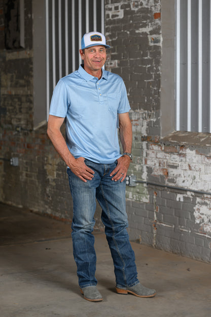 A man wearing a light blue polo shirt, blue jeans, gray suede boots, and a white cap with a brown logo, standing in front of a brick and corrugated metal wall.