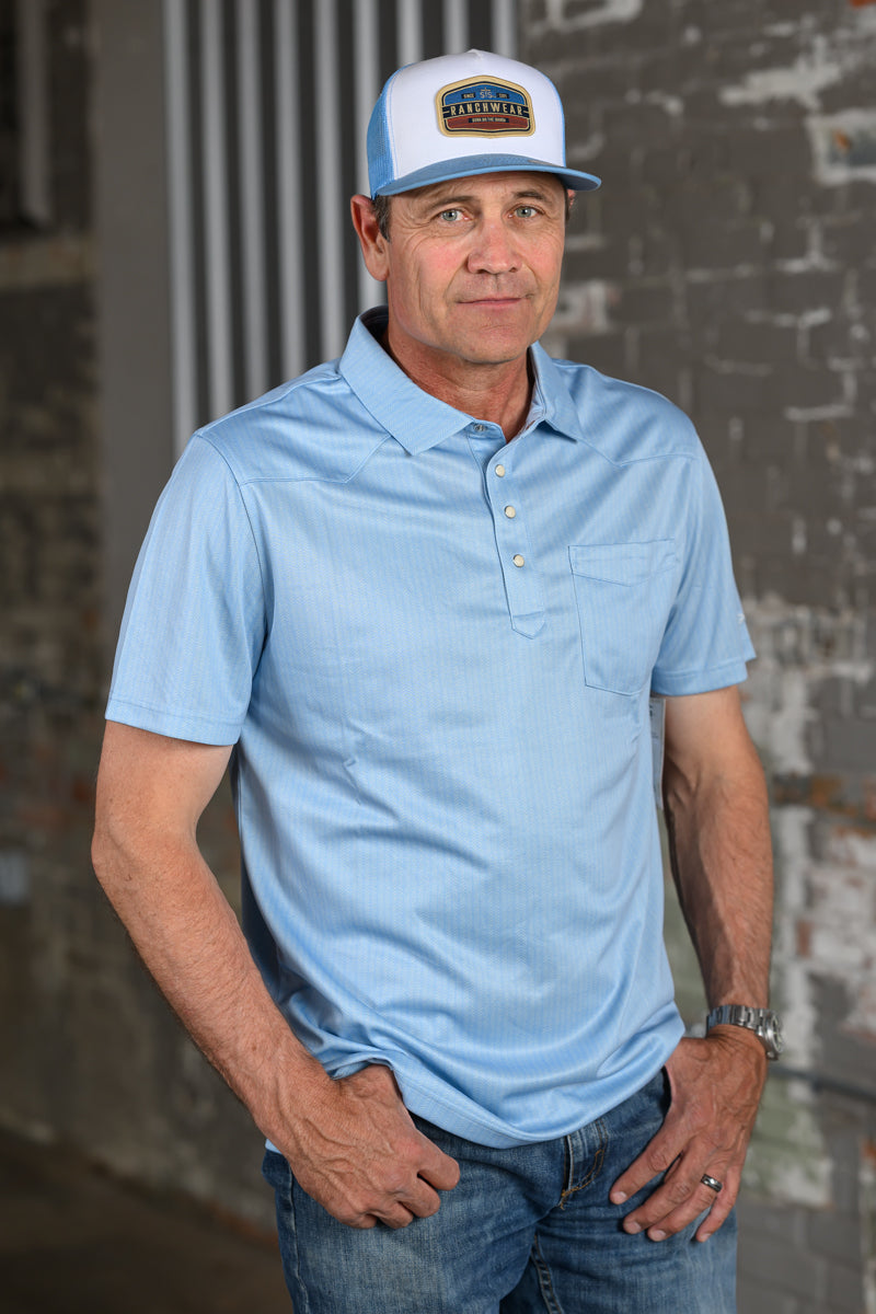 A man wearing a light blue short-sleeve polo shirt, blue jeans, and a white and blue St. Augustine baseball cap, standing against a rustic brick wall.