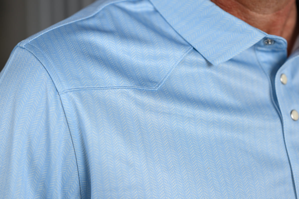 Close-up of a light blue textured button-down shirt on a model, showing the collar, shoulder seam, and white buttons.