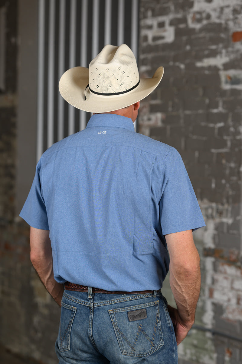 A man viewed from behind wearing a light blue short-sleeve button-up shirt, blue jeans, and a cream-colored cowboy hat with silver studs, standing against a rustic brick wall.