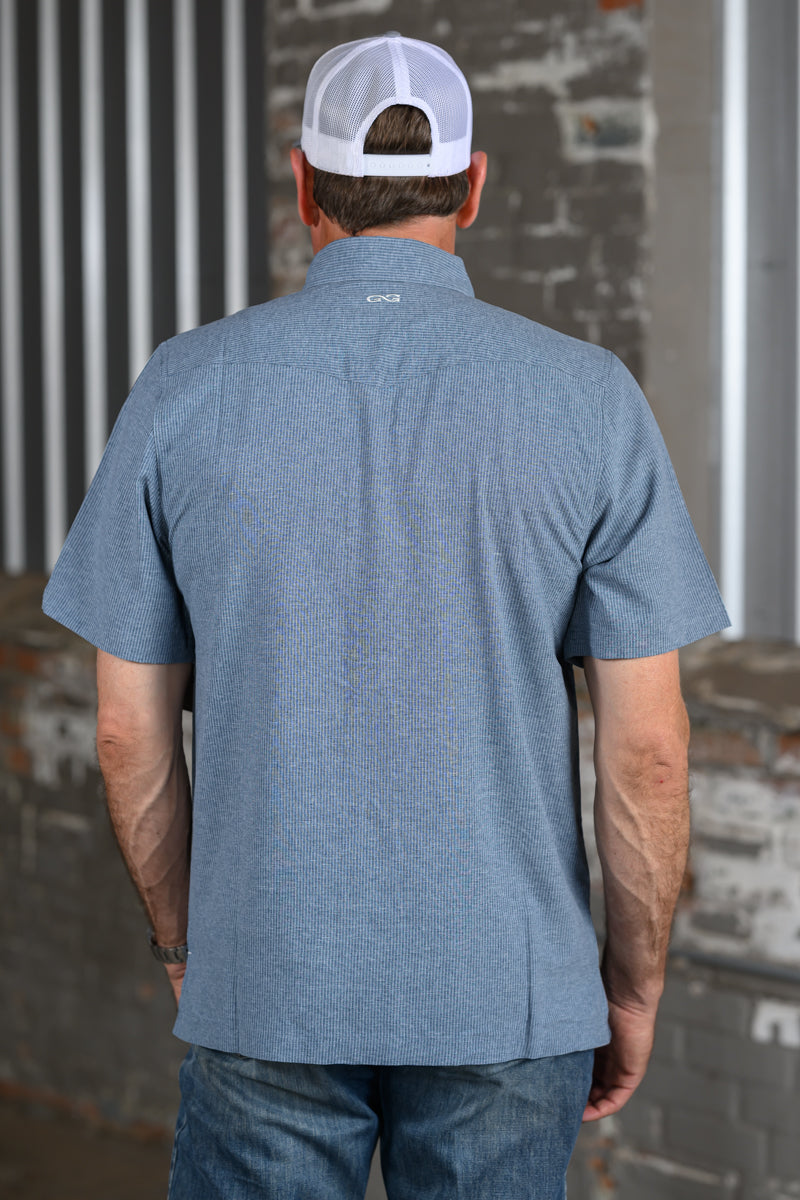 A man viewed from behind wearing a heathered blue short-sleeve button-up shirt and a white baseball cap, standing in front of a rustic brick and metal wall.