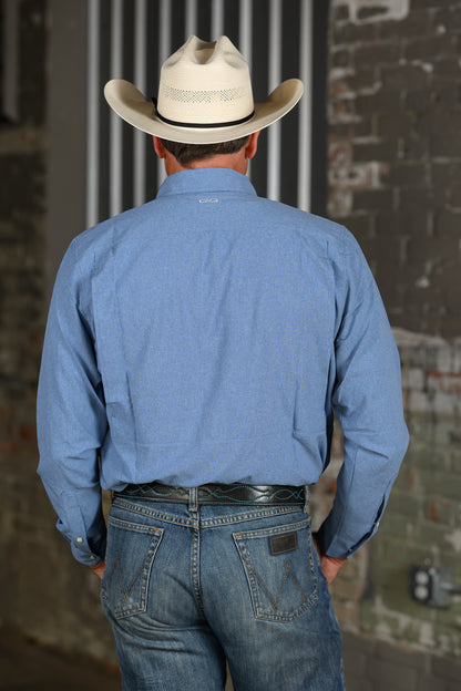A man viewed from behind wearing a light blue long-sleeve western shirt, blue jeans, a black belt, and a cream-colored cowboy hat, standing against a brick wall background.
