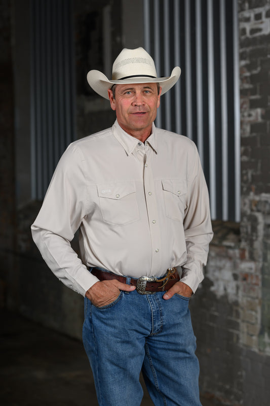A man wearing a light beige western long-sleeve button-up shirt, blue jeans, a brown leather belt, and a white cowboy hat, standing with hands in his pockets against a rustic industrial background.