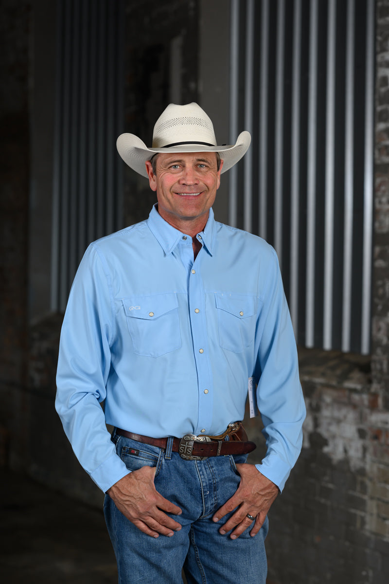 A smiling man wearing a light blue western-style button-down shirt, blue jeans, a brown belt, and a white cowboy hat, standing in front of a dark corrugated metal and brick wall.