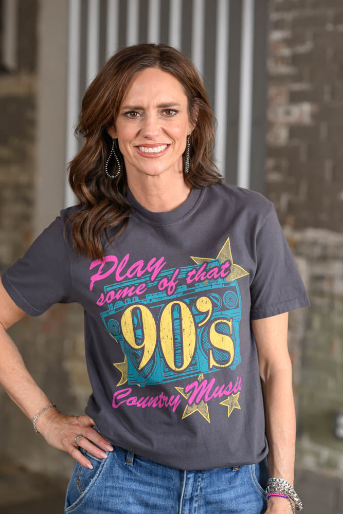 A woman wearing a dark gray graphic t-shirt with a colorful retro 90s design that reads 'Play some of that 90's Country Musix', paired with blue jeans, standing in front of a rustic brick and wood background.