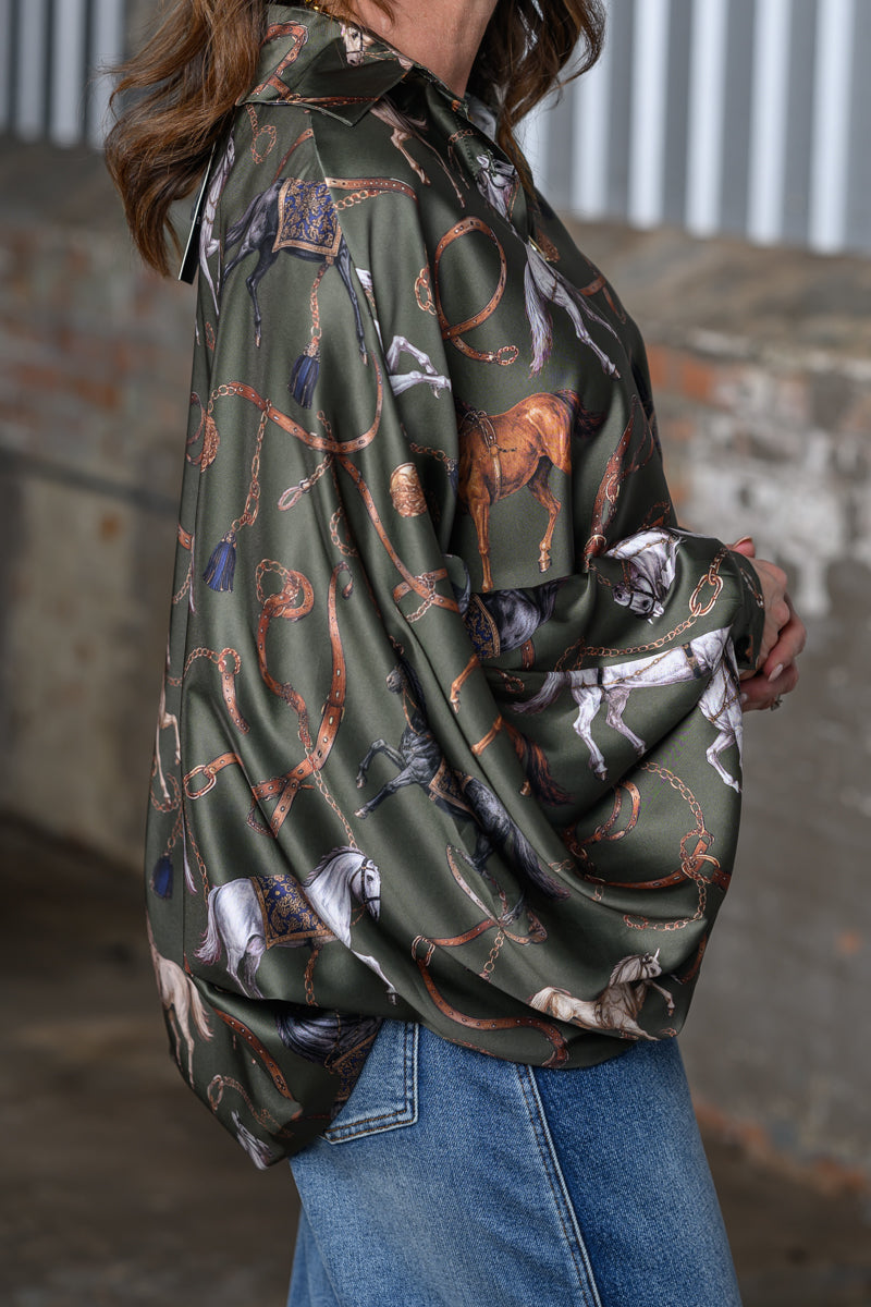 A woman wearing an olive green satin shirt with a western-themed print of horses, lasso ropes, and boots, paired with blue jeans.