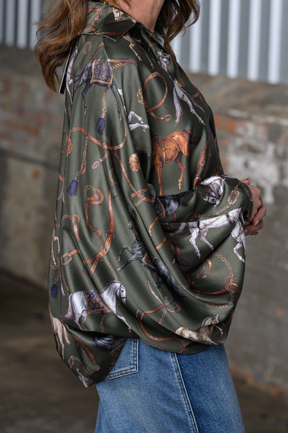 A woman wearing an olive green satin shirt with a western-themed print of horses, lasso ropes, and boots, paired with blue jeans.