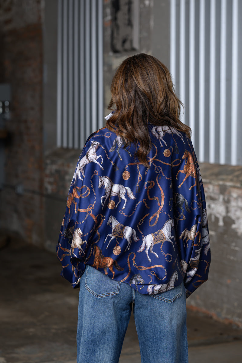 A woman viewed from behind wearing a navy blue satin bomber jacket with a white, brown, and gold horse and lasso print, paired with blue jeans in an industrial setting.