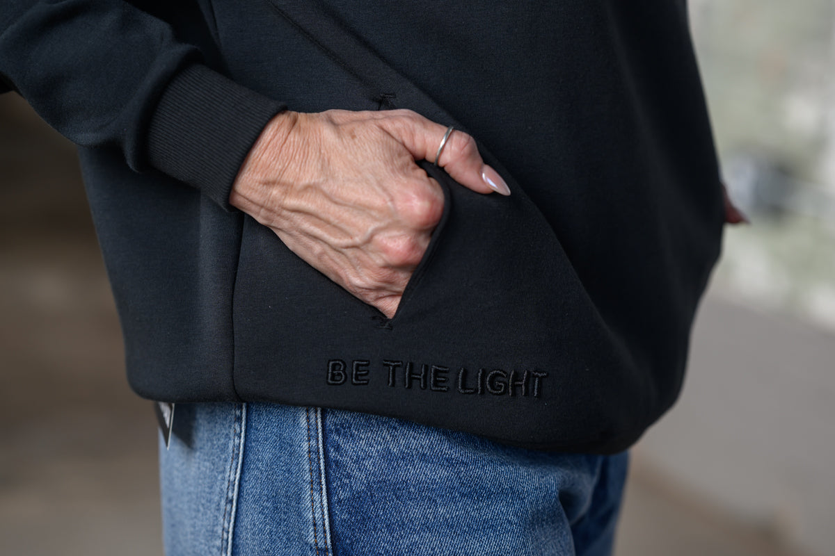 A close-up of a person wearing a black hoodie with a hand tucked into the front pocket. The hoodie features the text 'BE THE LIGHT' debossed on the lower pocket.